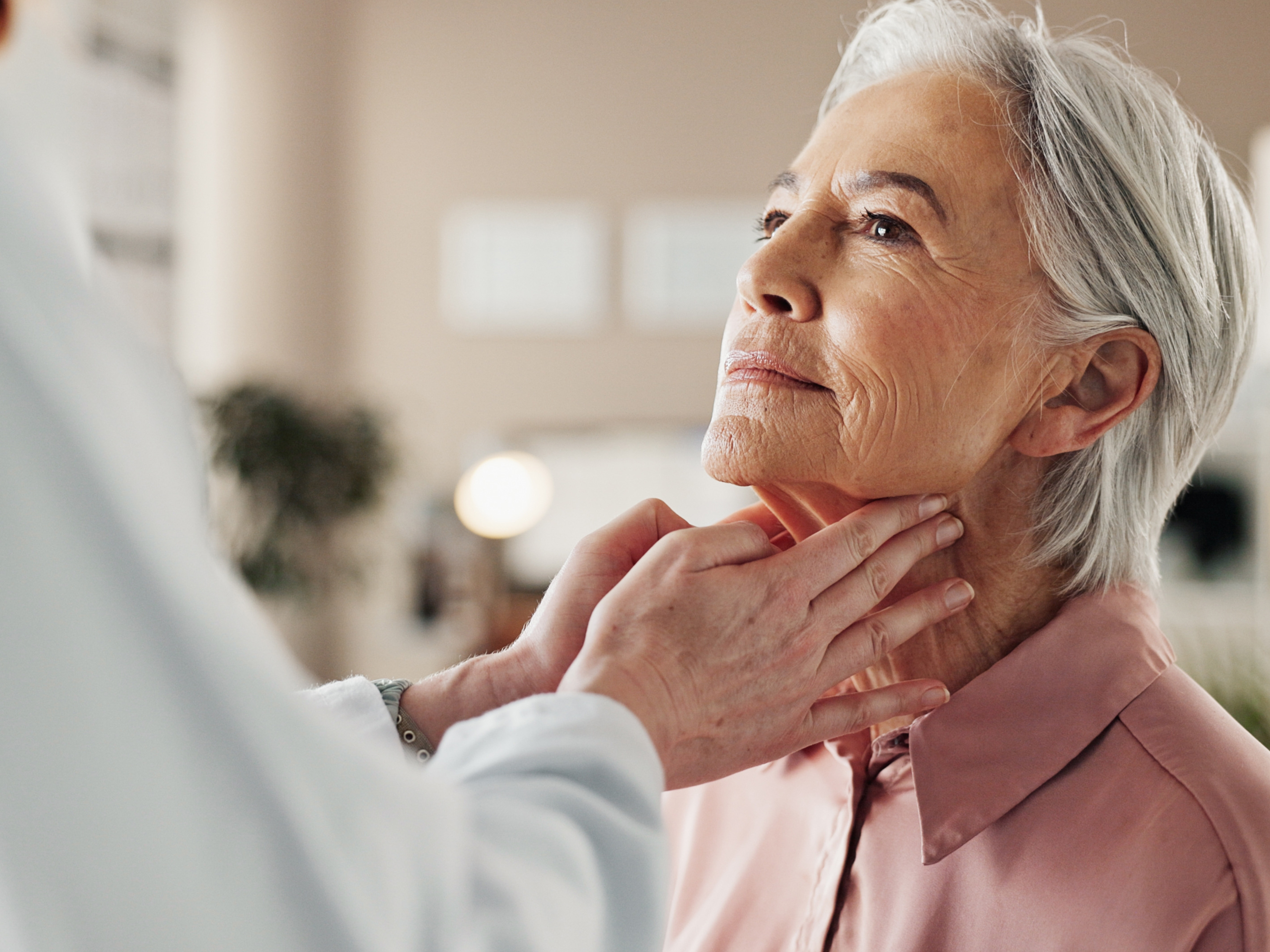 A older female wearing a pink shirt is having her throat examined by her primary care provider.